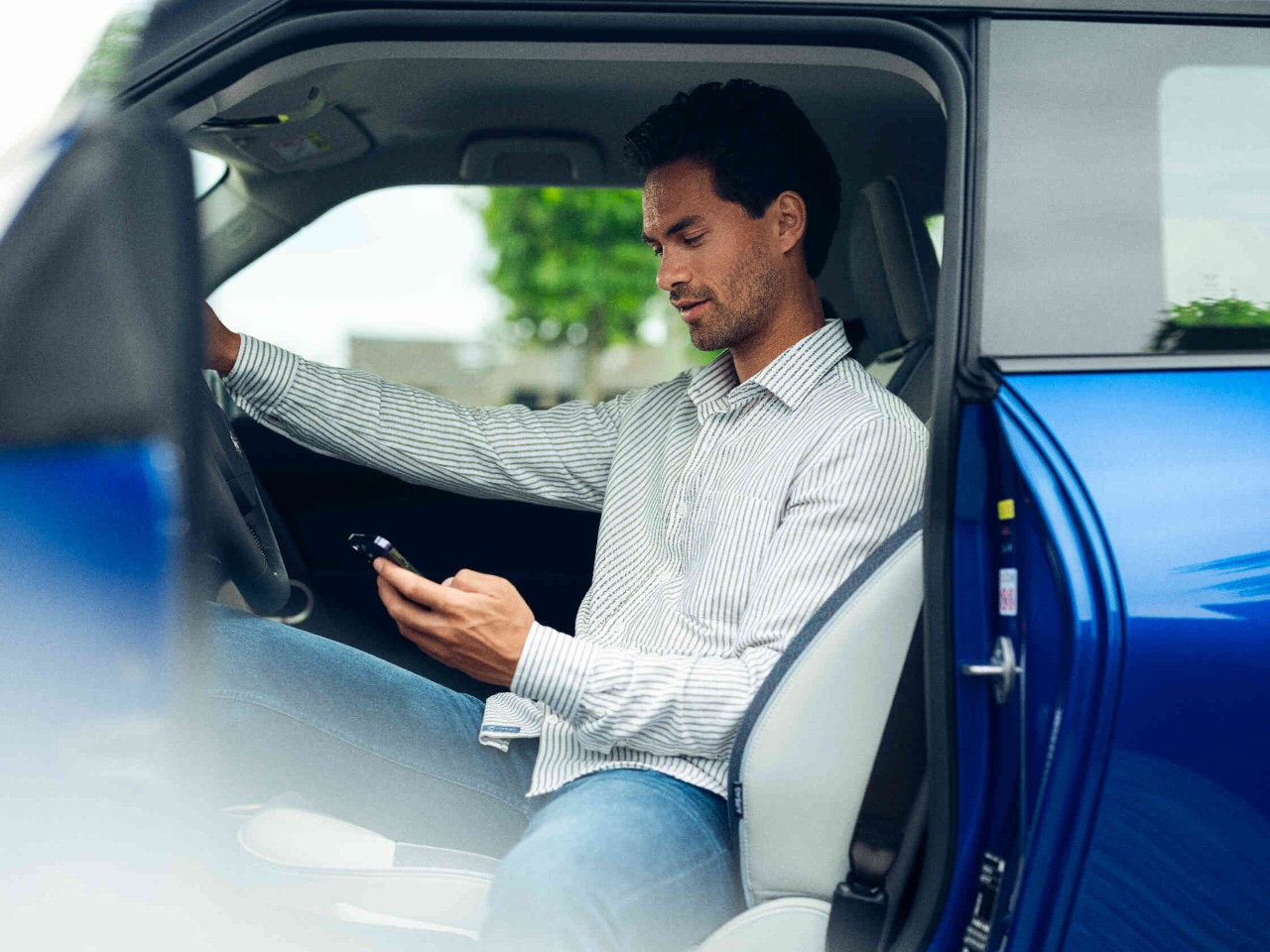Man-telefoon-bestuurdersstoel Man zit op de bestuurdersstoel van een stilstaande auto en kijkt op zijn telefoon de bestuurdersdeur staat open.